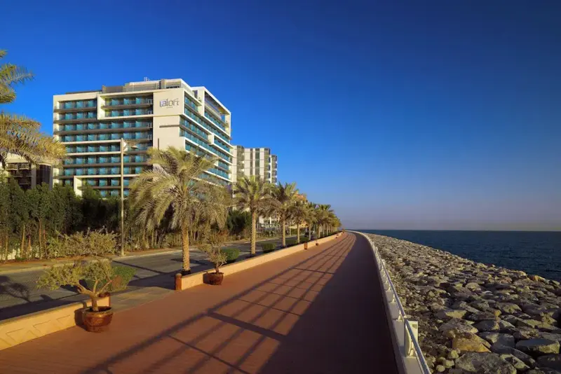 Vue d'un bâtiment moderne le long d'une promenade ensoleillée bordée de palmiers.