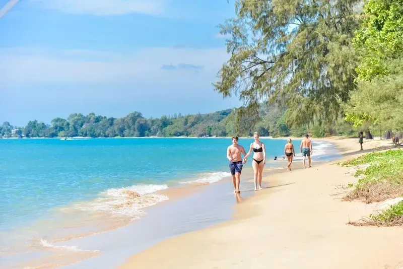 Des personnes se promènent sur une plage de sable fin bordée d'arbres.