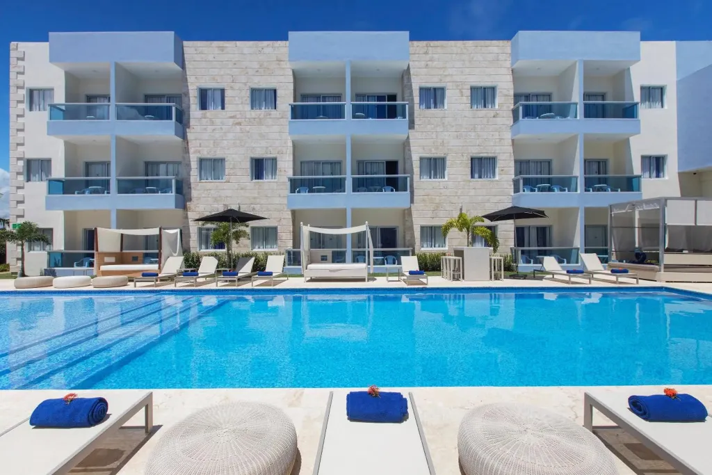 Une piscine turquoise bordée de transats blancs invite à la détente devant un bâtiment moderne.