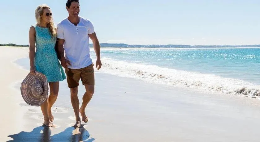 Un couple marche pieds nus le long d'une plage de sable fin, bordée par une mer turquoise.