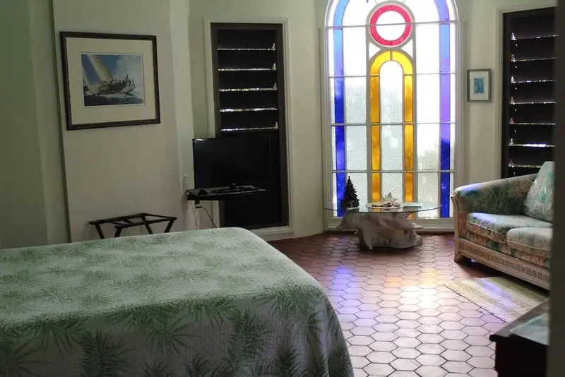 Une chambre lumineuse avec un lit recouvert d'une couverture verte et un fauteuil fleuri près d'une fenêtre colorée.