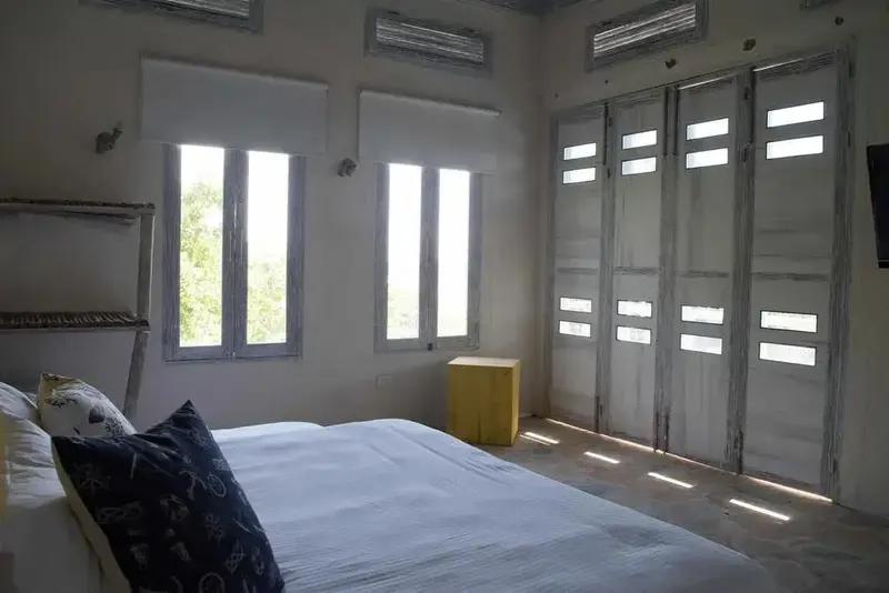 Une chambre lumineuse avec un lit recouvert d'un drap blanc et des ouvertures sur un extérieur verdoyant.