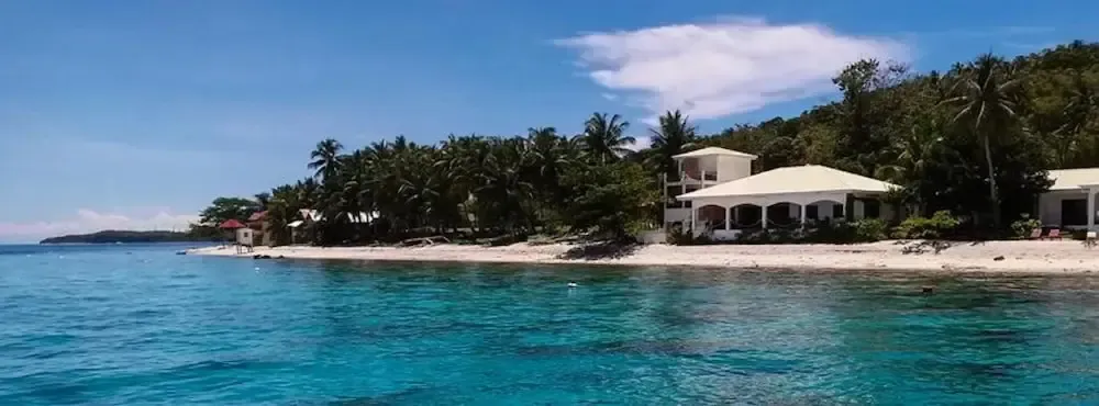 Vue d'une plage de sable blanc bordée de végétation luxuriante et de bâtiments clairs.