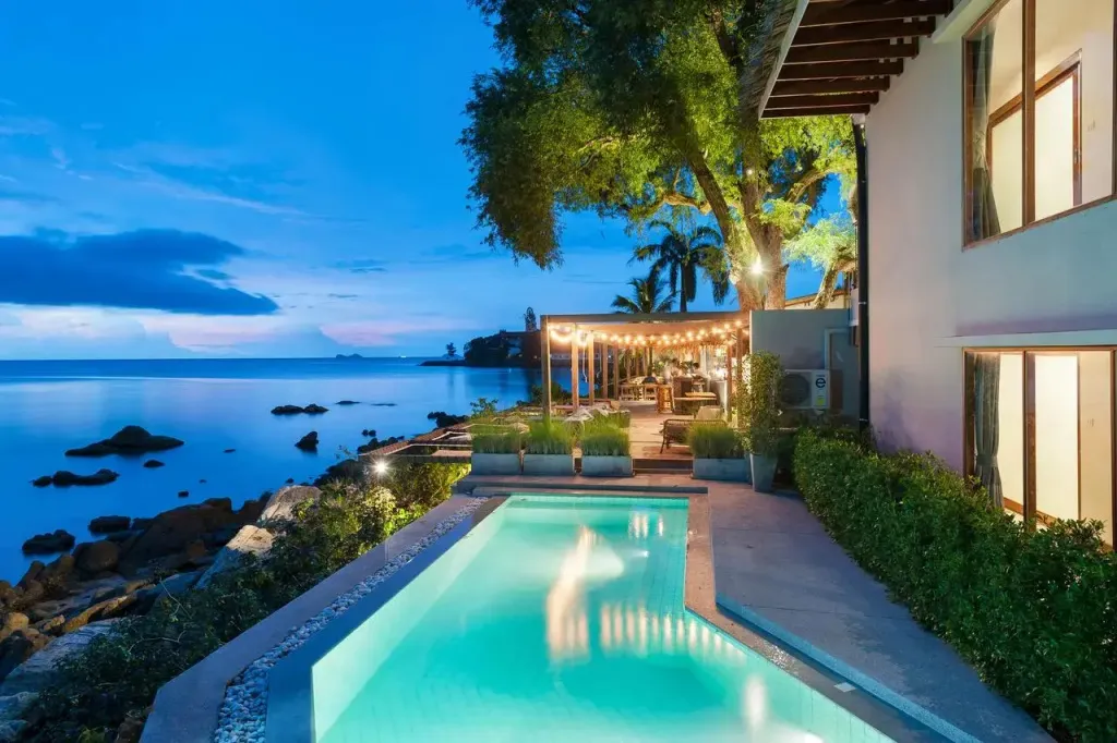 Une piscine rectangulaire illuminée borde un espace extérieur avec une salle à manger et une vue sur la mer.