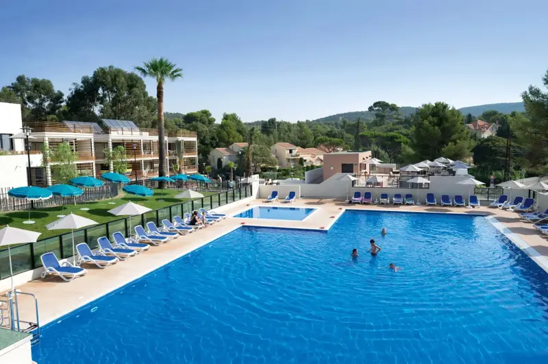 Une piscine rectangulaire bordée de transats sous des parasols, avec des bâtiments en arrière-plan.
