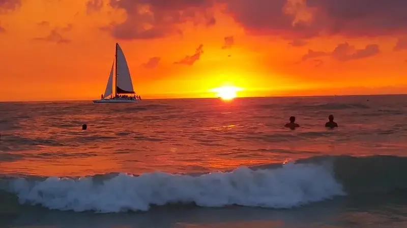 Le soleil se couche sur l'océan, illuminant les vagues et un voilier à l'horizon.