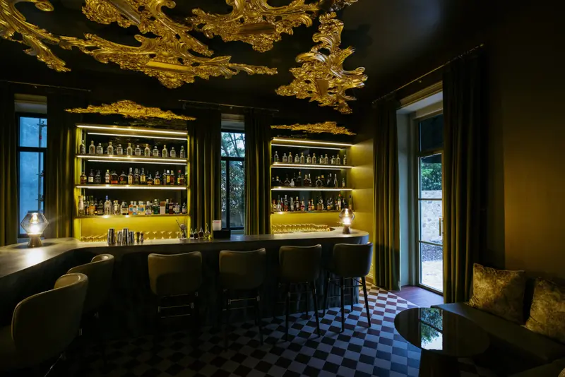 Un comptoir de bar doré est éclairé par des lampes, avec des tabourets et des bouteilles exposées sur des étagères.