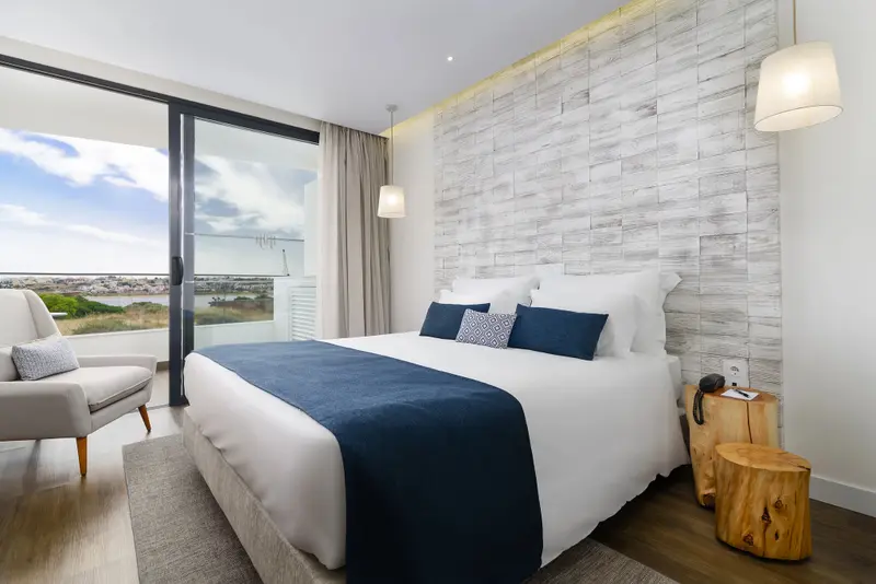 Une chambre lumineuse avec un grand lit et un fauteuil près d'une baie vitrée.