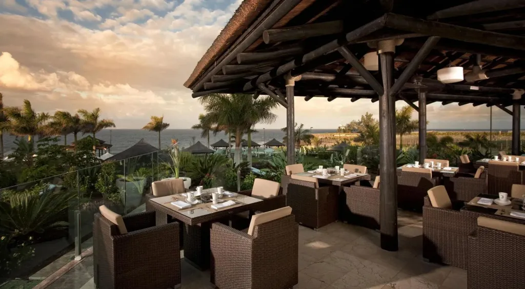 Terrasse ombragée avec tables et chaises pour les repas, vue sur la mer au loin.