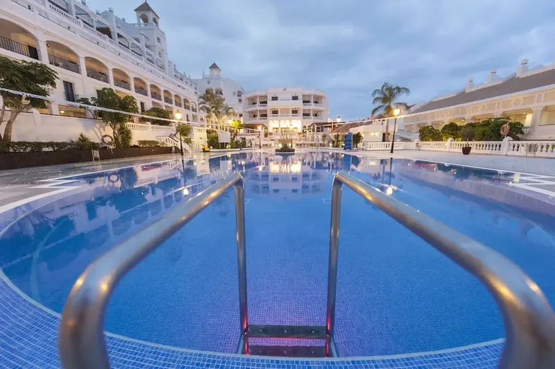 Une piscine rectangulaire avec des escaliers en inox, entourée de bâtiments illuminés.