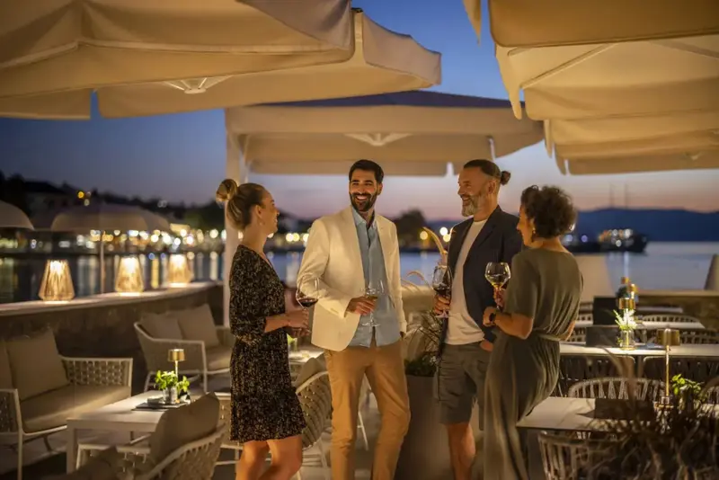 Quatre personnes discutent autour de verres sur une terrasse ombragée en soirée.