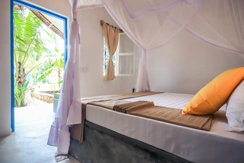 Une chambre lumineuse avec un lit recouvert d'une moustiquaire et une porte ouverte sur une végétation luxuriante.
