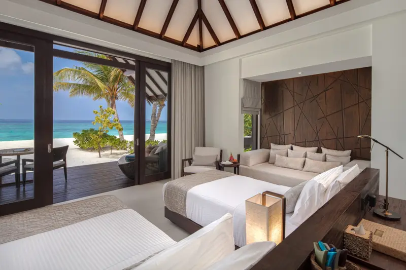 Une chambre lumineuse s'ouvre sur une terrasse en bois donnant sur une plage de sable blanc et une mer turquoise.