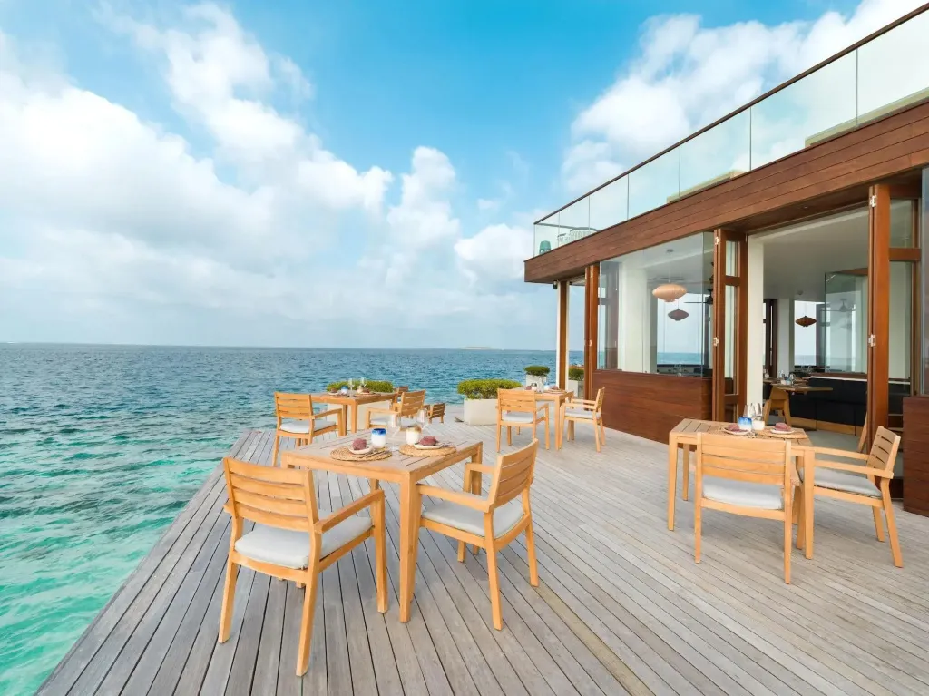 Tables de restaurant en bois clair sur une terrasse avec vue sur la mer.