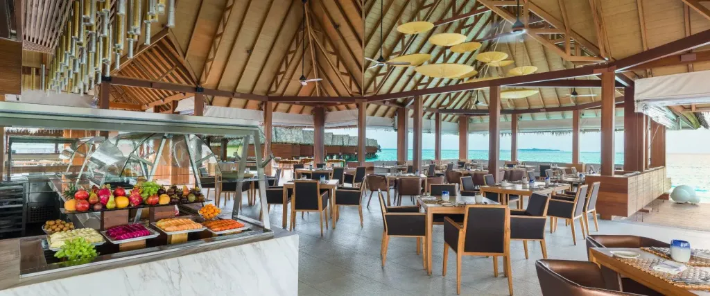 Une salle lumineuse avec un buffet de fruits et des tables de repas face à l'océan.