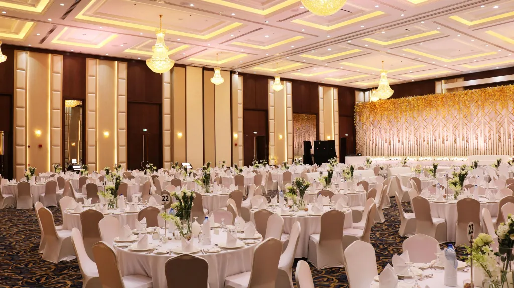Une grande salle de réception est mise en scène pour un banquet avec des tables rondes nappées et décorées de fleurs.