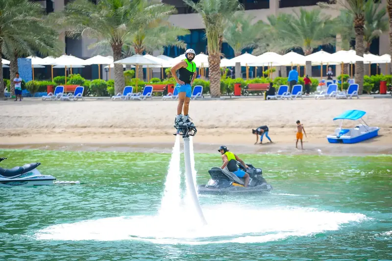 Une personne pratique le flyboard au-dessus de l'eau turquoise, avec une plage en arrière-plan.