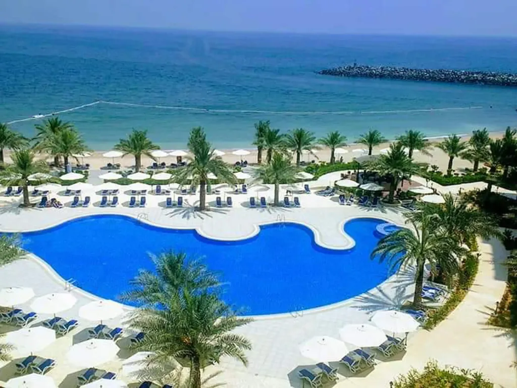 Piscine sinueuse bordée de transats et de palmiers, avec une plage en arrière-plan.