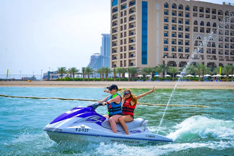 Deux personnes s'amusent sur un jet ski en mer devant un grand bâtiment.