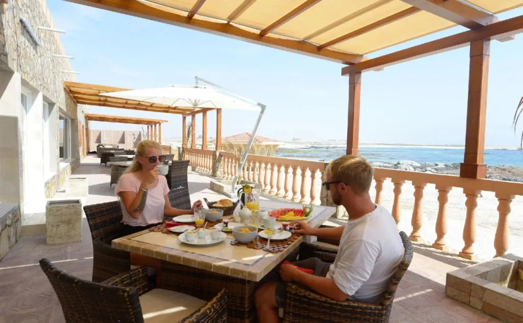 Un couple prend son petit-déjeuner à une table sur une terrasse ombragée avec vue sur la mer.