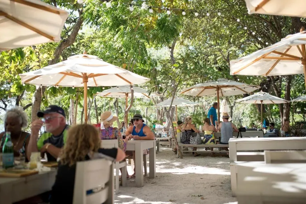 Des convives déjeunent à l'ombre de parasols sur une terrasse ensoleillée.