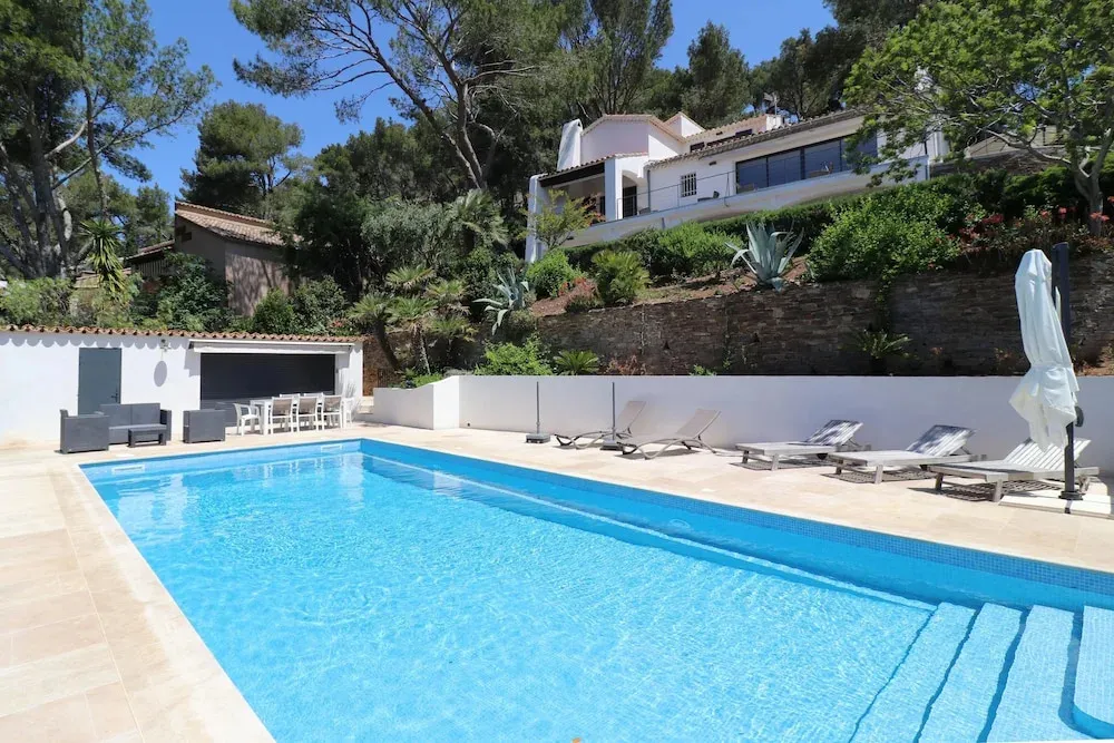 Une piscine rectangulaire turquoise est bordée de transats et d'un parasol blanc.