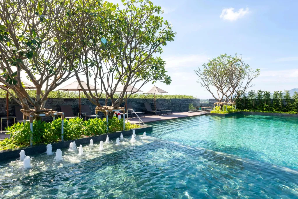 Une piscine rectangulaire avec des jets d'eau et des transats à l'ombre de parasols.