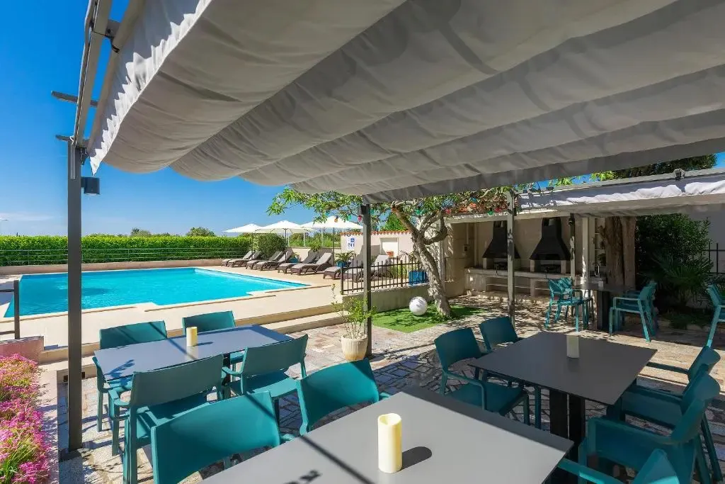 Tables et chaises turquoise sous une pergola, avec une piscine en arrière-plan.