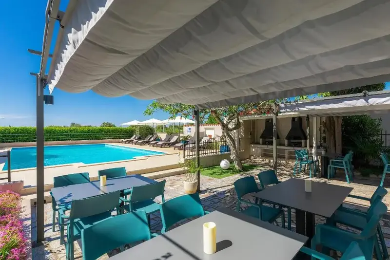 Tables et chaises turquoise sous une pergola, avec une piscine en arrière-plan.