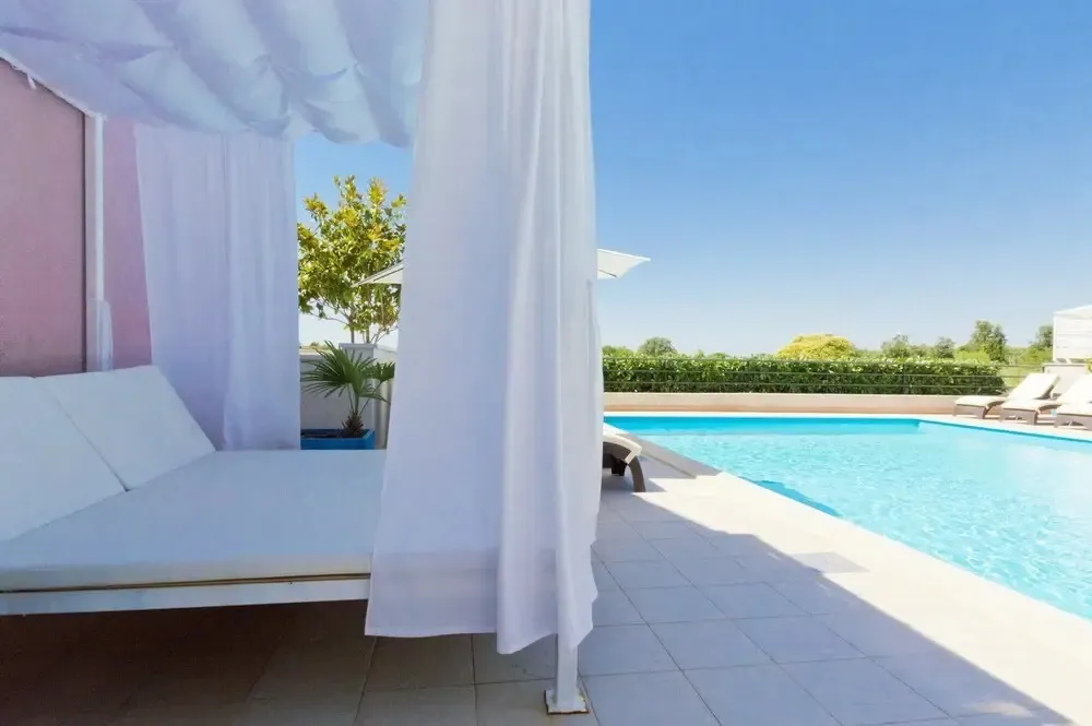 Transats blancs sous des rideaux vaporeux au bord d'une piscine ensoleillée.