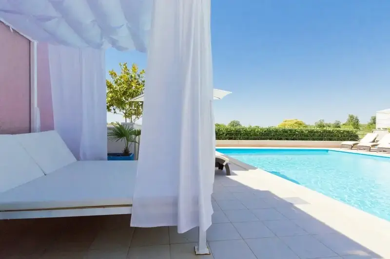 Transats blancs sous des rideaux vaporeux au bord d'une piscine ensoleillée.