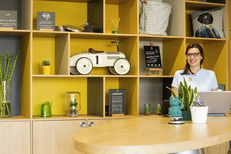 Une employée souriante se tient derrière un comptoir en bois dans un espace lumineux et décoré.