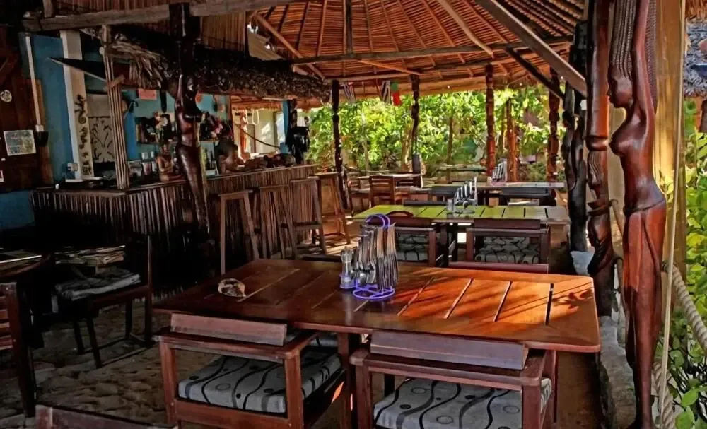 Une salle à manger en bois avec des tables et des chaises est éclairée par la lumière du soleil.