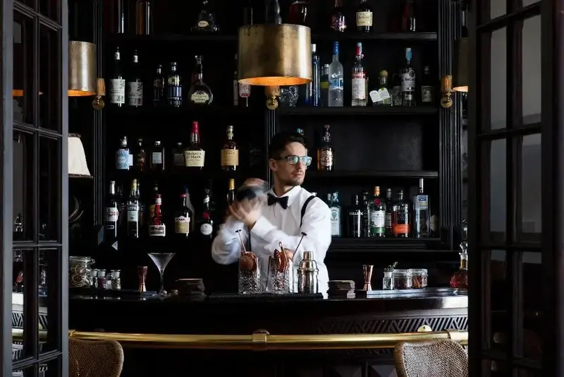 Un barman prépare un cocktail derrière un comptoir richement garni de bouteilles.