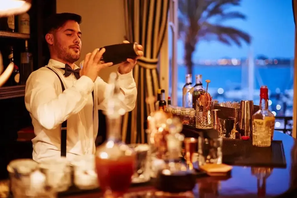 Un barman prépare un cocktail derrière un comptoir bien garni, avec vue sur la mer.