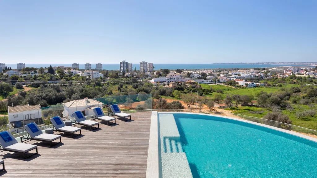 Terrasse en bois avec transats bleus dominant une piscine et une vue sur la mer.