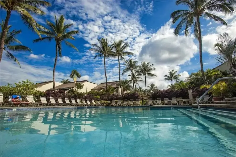 Une piscine turquoise bordée de chaises longues sous des palmiers au ciel nuageux.