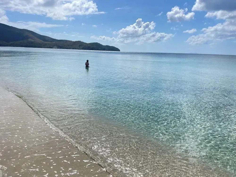 Une personne se tient dans l'eau turquoise le long d'une plage de sable clair.