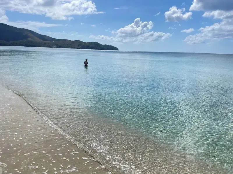 Une personne se tient dans l'eau turquoise le long d'une plage de sable clair.