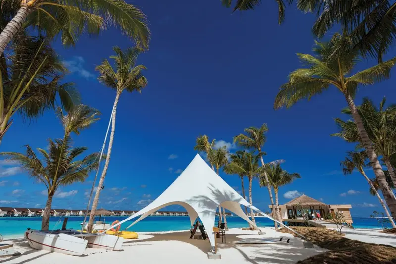Tonnelle blanche sur une plage de sable fin bordée de palmiers sous un ciel bleu.