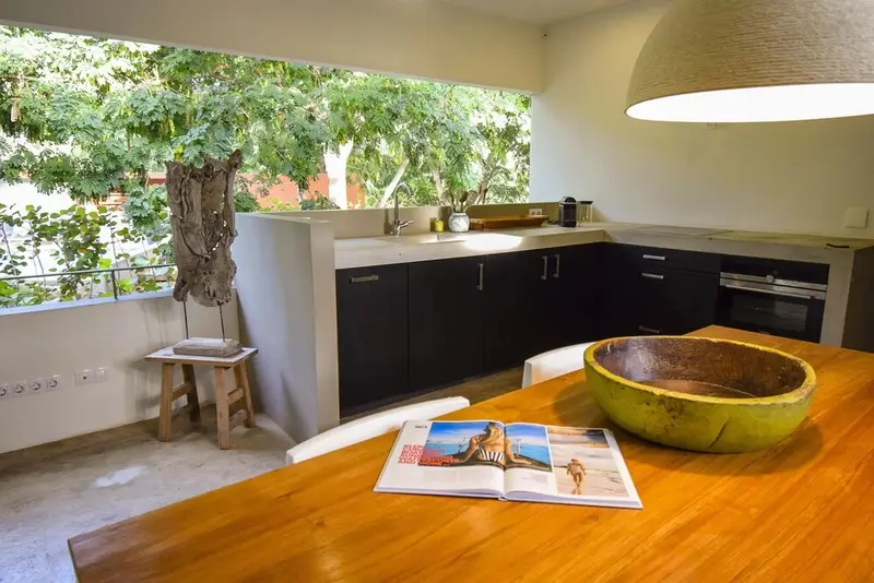 Une table en bois est mise en scène devant une cuisine équipée et une fenêtre ouverte sur la végétation.
