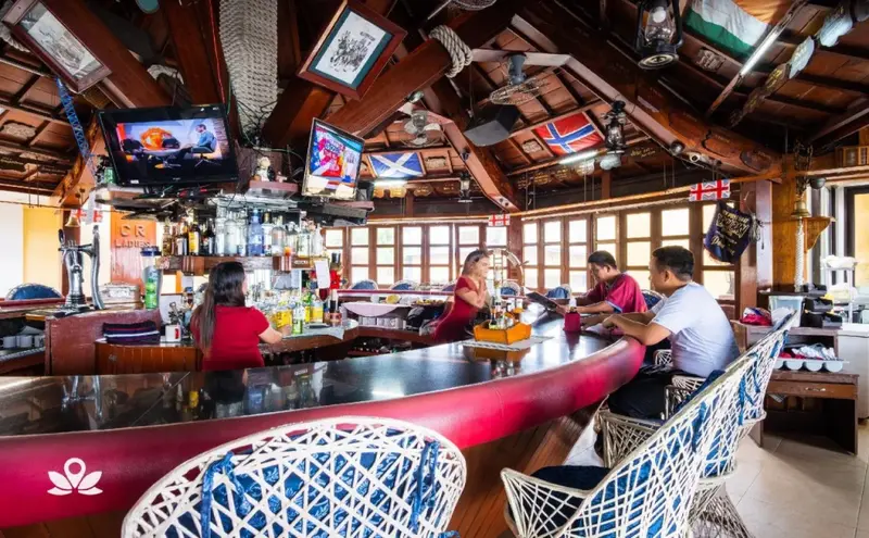 Un comptoir de bar en bois rouge accueille des clients dans une ambiance tropicale.