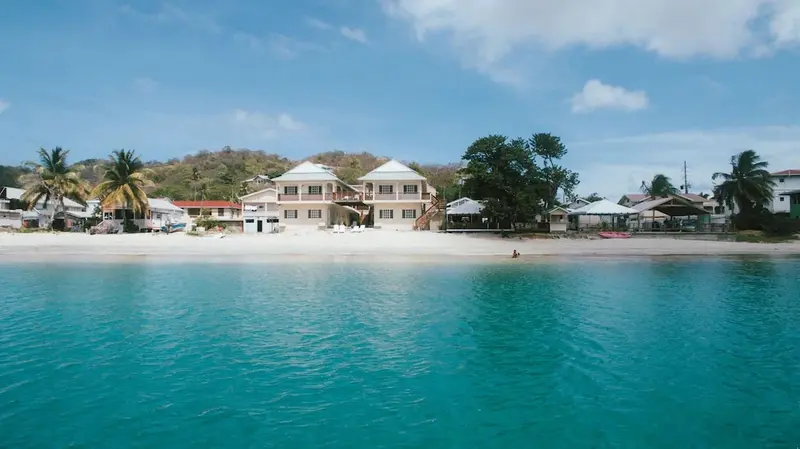 Vue d'une plage de sable blanc bordée de bâtiments et de végétation tropicale sous un ciel bleu.