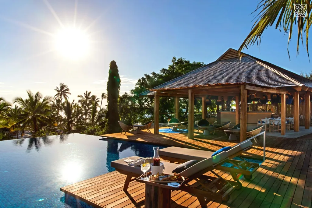 Transats et table basse avec boissons au bord d'une piscine à débordement ensoleillée.