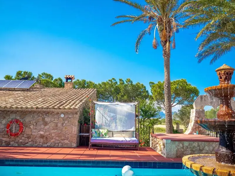 Une fontaine orne le bord d'une piscine turquoise, avec un espace de détente ombragé à proximité.