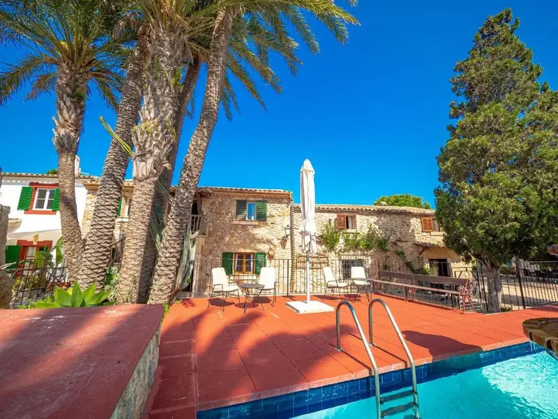 Une piscine bordée d'une terrasse en bois rouge, avec des transats et un parasol près d'un bâtiment en pierre.