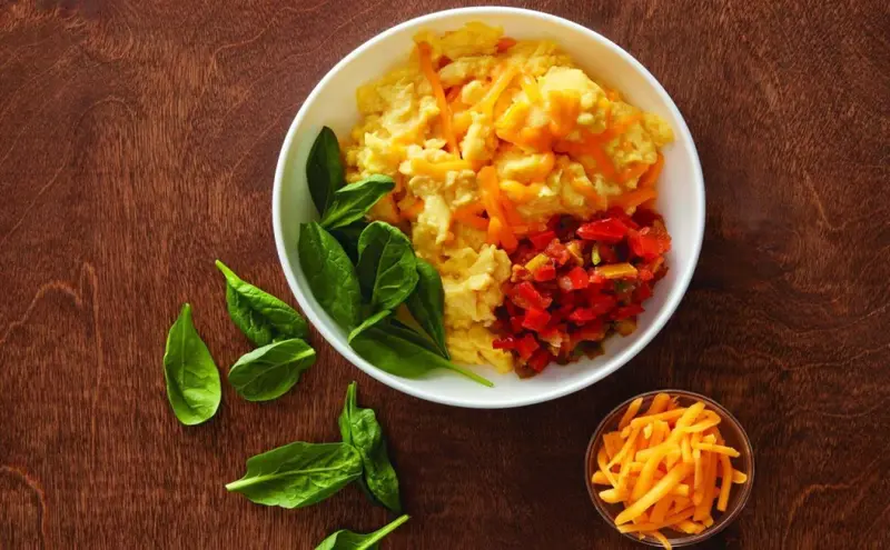 Un bol d'œufs brouillés garnis de légumes et de fromage, accompagné d'épinards frais et d'un ramequin de fromage râpé.