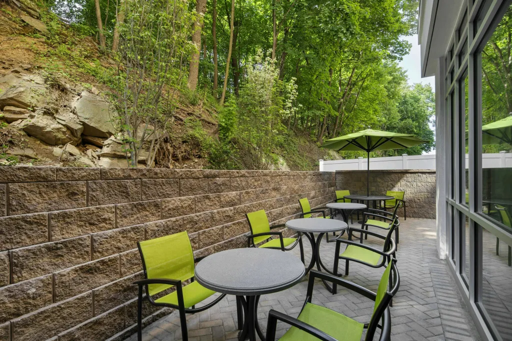 Terrasse ombragée avec tables et chaises vertes près d'un mur en pierre.