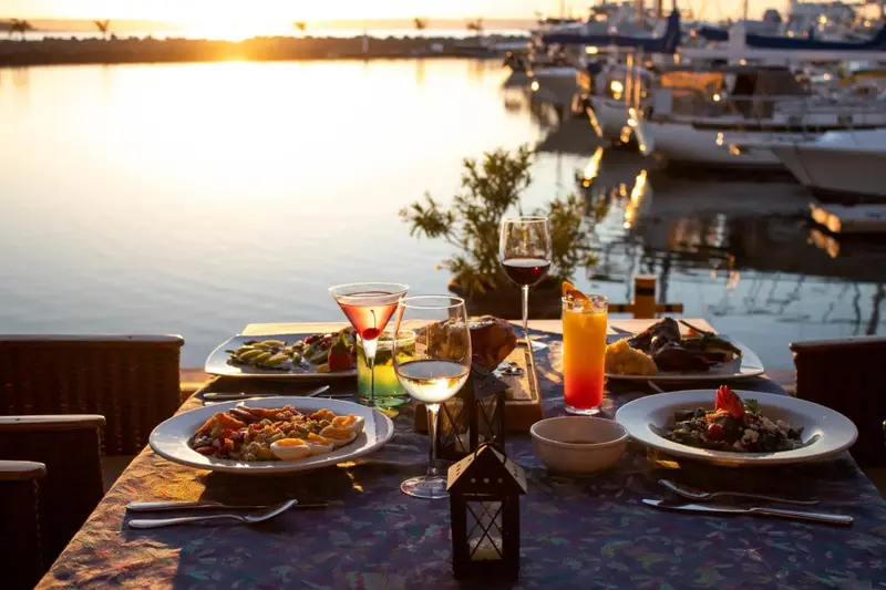 Une table dressée avec des plats et des boissons face à un port de plaisance au coucher du soleil.