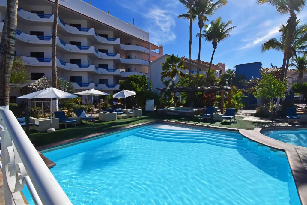 Piscine turquoise bordée de transats sous des parasols blancs, avec un bâtiment aux balcons fleuris en arrière-plan.